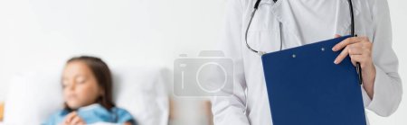 Photo for Doctor holding clipboard near blurred child on hospital ward, banner - Royalty Free Image