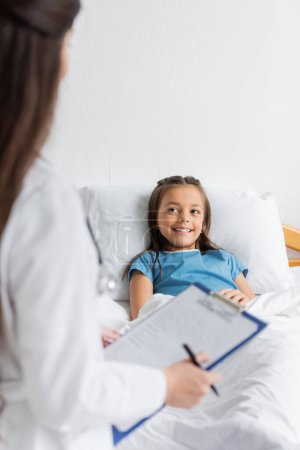 Téléchargez les photos : Enfant souriant allongé sur le lit tandis que le médecin flou tenant presse-papiers dans la salle d'hôpital - en image libre de droit