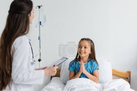 Positive girl doing praying hands gesture near blurred doctor with clipboard in clinic 