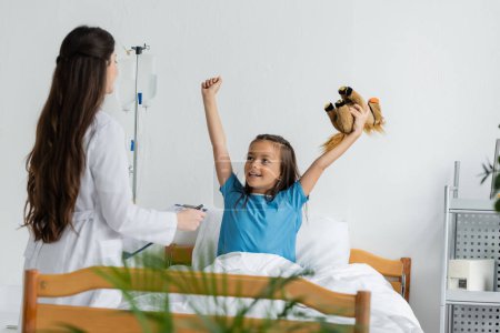 Kind mit Spielzeug zeigt Ja-Geste in der Nähe von Kinderarzt mit Klemmbrett auf Krankenhausstation 