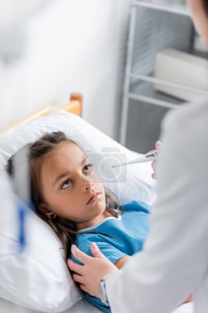Doctor holding thermometer and touching child on clinic bed 