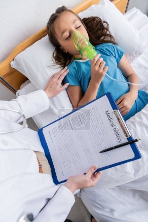 Kinderarzt hält Klemmbrett in der Nähe von Kind mit Sauerstoffmaske auf Bett in Klinik 