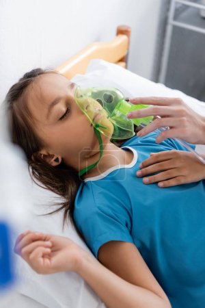 Arzt mit Sauerstoffmaske nahe Kind auf Bett im Krankenhaus 