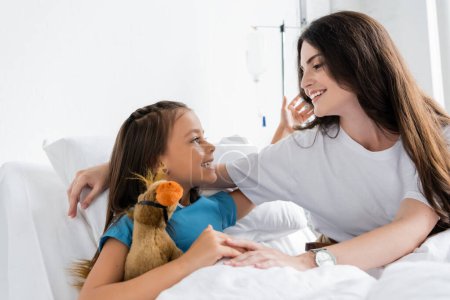 Positive kid hugging mother and holding soft toy on bed in clinic 