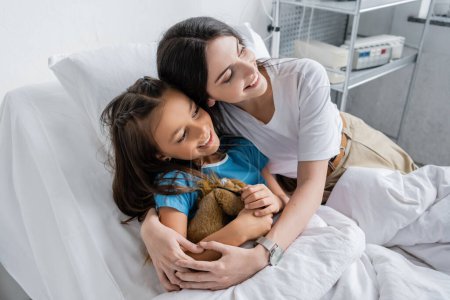 Happy parent hugging daughter with toy on bed in clinic 