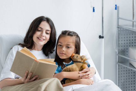 Smiling woman reading book near daughter with toy on bed in hospital bed 