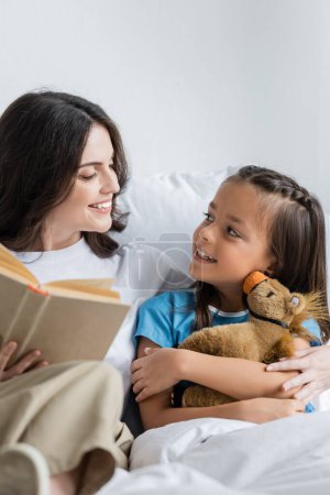 Mother holding book near smiling child with toy on clinic bed 