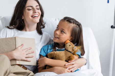 Cheerful woman holding book near daughter with soft toy in hospital 