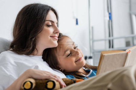 Positive woman holding soft toy while reading book with mother in hospital ward 