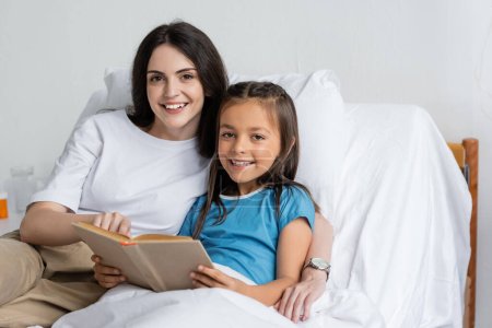 Smiling woman and daughter looking at camera near book on bed in hospital 