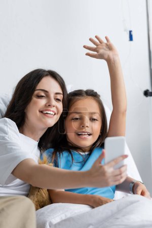 Cheerful woman holding smartphone near daughter during video call in clinic 