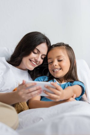 Mujer sonriente e hija sosteniendo teléfono inteligente en la cama en la sala de hospital 