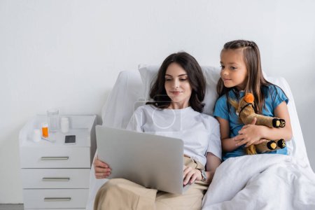 Mutter hält Laptop neben Tochter im Patientenmantel auf Krankenhausstation 