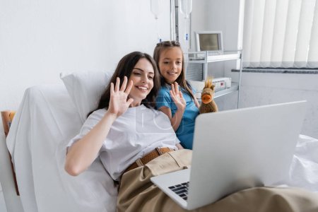 Lächelnde Frau und Mädchen im Patientenkleid mit Videoanruf auf Laptop auf Krankenhausstation 