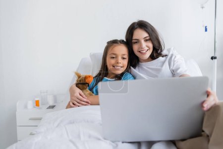 Lächelnde Eltern halten Laptop in der Hand und umarmen Tochter im Patientenkleid auf Krankenhausstation 