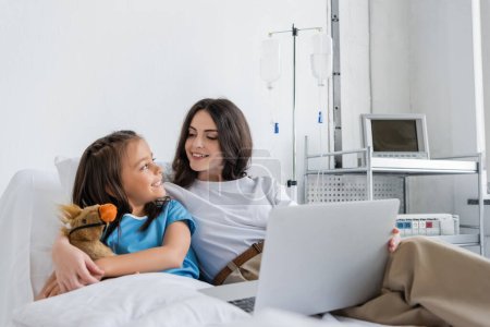 Child in patient gown smiling to mother with laptop on bed in hospital 