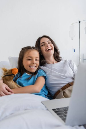 Glückliches Kind im Patientenkleid mit Blick auf Laptop neben Mutter und Stofftier auf Bett im Krankenhaus 