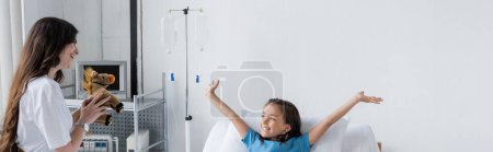 Parent holding soft toy near cheerful daughter in patient gown in clinic, banner 