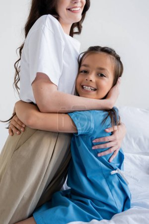 Parent hugging smiling daughter in patient gown on bed in hospital bed 
