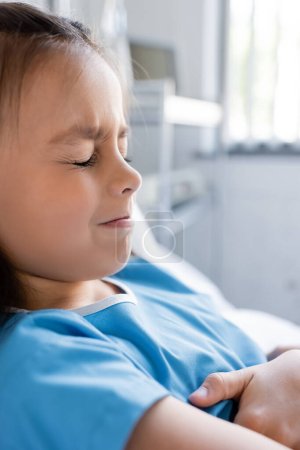 Téléchargez les photos : Enfant mécontent et malade en robe de patient couché sur le lit à la clinique - en image libre de droit