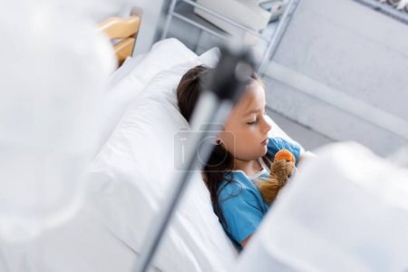 Blick aus der Vogelperspektive auf krankes Kind mit Spielzeug, das auf Bett in Klinik liegt 
