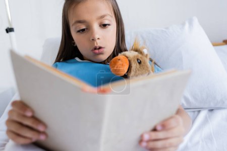 Kid in patient gown reading book near toy on hospital bed 