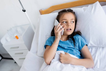 Ill child talking on smartphone while lying on bed in clinic 