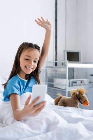 Positive kid in patient gown having video call on smartphone near soft toy in bed in clinic 