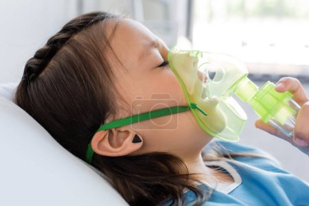 Diseased kid holding oxygen mask while lying on bed in clinic 