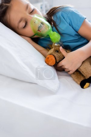 Ill girl in patient gown and oxygen mask holding toy on bed in hospital 