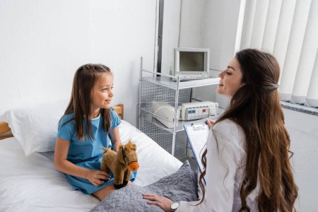 Lächelnder Arzt hält Klemmbrett und spricht mit Kind mit Spielzeug auf Bett im Krankenhaus 