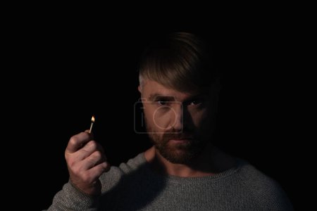 Foto de Hombre en la oscuridad sosteniendo partido iluminado y mirando a la cámara durante apagón eléctrico aislado en negro - Imagen libre de derechos