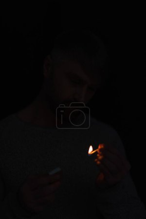 man with matchbox looking at burning match in darkness isolated on black
