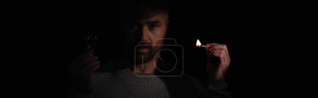 Foto de Hombre con bombilla y fósforo iluminado mirando a la cámara durante el apagón de energía aislado en negro, pancarta - Imagen libre de derechos