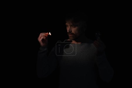 man in darkness holding electric bulb and looking at flame of lit match isolated on black