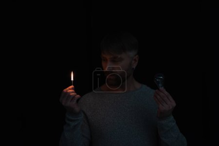man looking at flame of lit match while holding electric bulb isolated on black