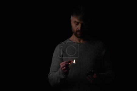 Mann in der Dunkelheit hält Streichholzschachtel in der Hand und betrachtet beleuchtetes Streichholz isoliert auf schwarz