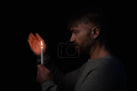 Foto de Vista lateral del hombre sosteniendo la mano cerca de la llama de la vela ardiente aislada en negro - Imagen libre de derechos