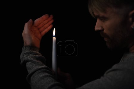Téléchargez les photos : Homme regardant la flamme de la bougie allumée pendant une panne de courant isolé sur noir - en image libre de droit
