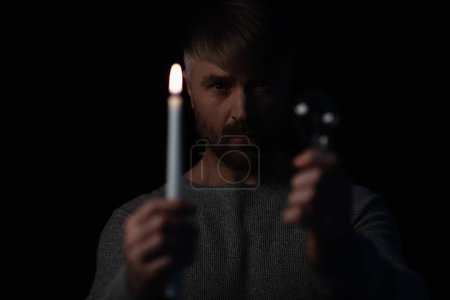 Photo for Man looking at camera while holding lit candle and light bulb isolated on black - Royalty Free Image