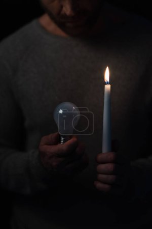 przycięty widok człowieka w ciemności trzymając zapaloną świecę i żarówkę elektryczną odizolowaną na czarno