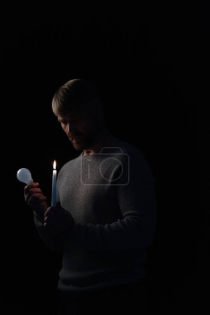 Erwachsener Mann mit Glühbirne und brennender Kerze während Stromabschaltung isoliert auf schwarz