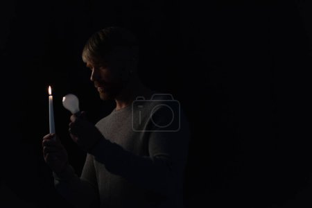 hombre sosteniendo la bombilla y la vela encendida en la oscuridad aislado en negro