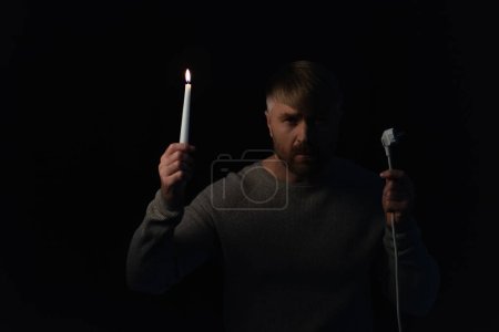 Foto de Hombre con enchufe y vela encendida mirando a la cámara durante el apagado eléctrico aislado en negro - Imagen libre de derechos