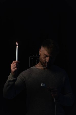 man with lit candle looking at electric plug during energy blackout isolated on black