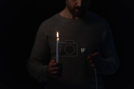 częściowy widok człowieka patrzącego na wtyczkę elektryczną trzymającego świecę podczas zaniku zasilania izolowanego na czarno