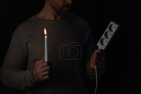 vista parcial del hombre sosteniendo extensor de enchufe y vela encendida durante corte de energía aislado en negro