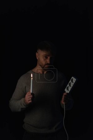 Mann in Dunkelheit durch Stromausfall im Stehen mit brennender Kerze und Steckdosenverlängerung isoliert auf schwarz