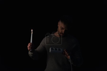 man with lit candle looking at  socket extender during power outage isolated on black