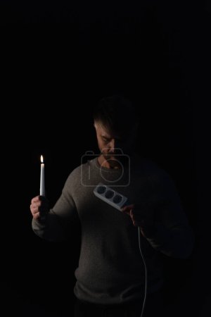 Erwachsener Mann mit brennender Kerze in der Dunkelheit und Blick auf Steckdosenverlängerer isoliert auf schwarz